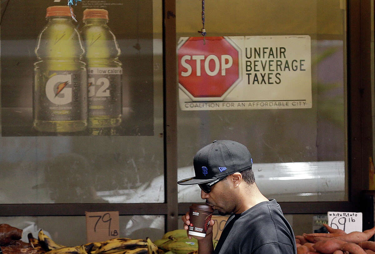 In this July 7, 2014 photo, a man drinks out of a coffee cup as he walks under a sign opposed to taxes on sugary drinks posted in the window of Casa Thai Market in San Francisco. San Francisco and Berkeley are aiming to become the first U.S. cities to pass per-ounce taxes on sugary drinks. (AP Photo/Jeff Chiu) ORG XMIT: CAJC306