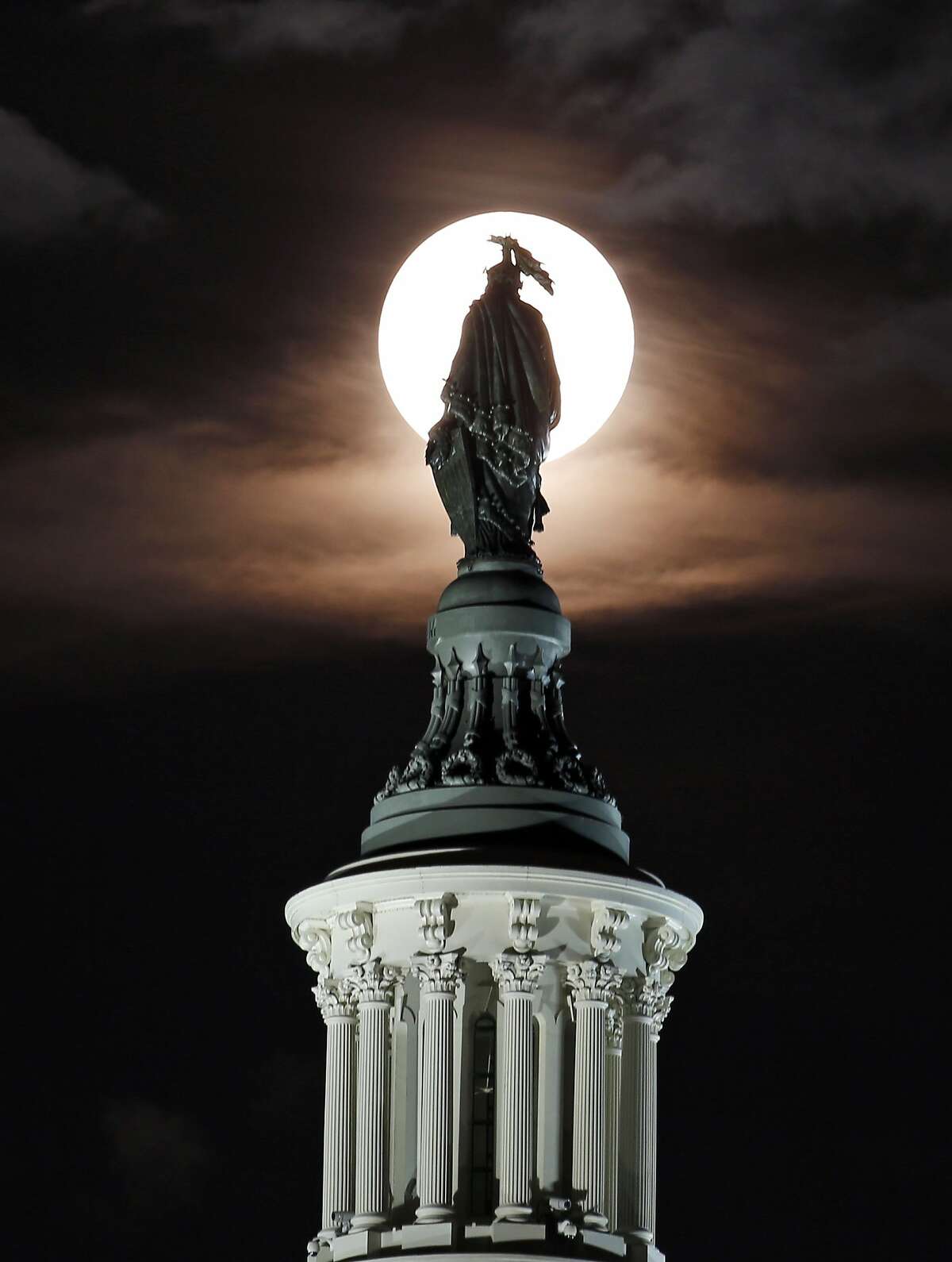 The supermoon rises through the clouds behind the bronze Statue of Freedom by Thomas Crawford atop the U.S. Capitol.