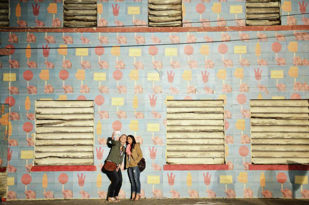 Kendra Erskine (left) and Cassie Rubio take a selfie next to a wall with Luminaria's design pasted on it near the Tobin Center.