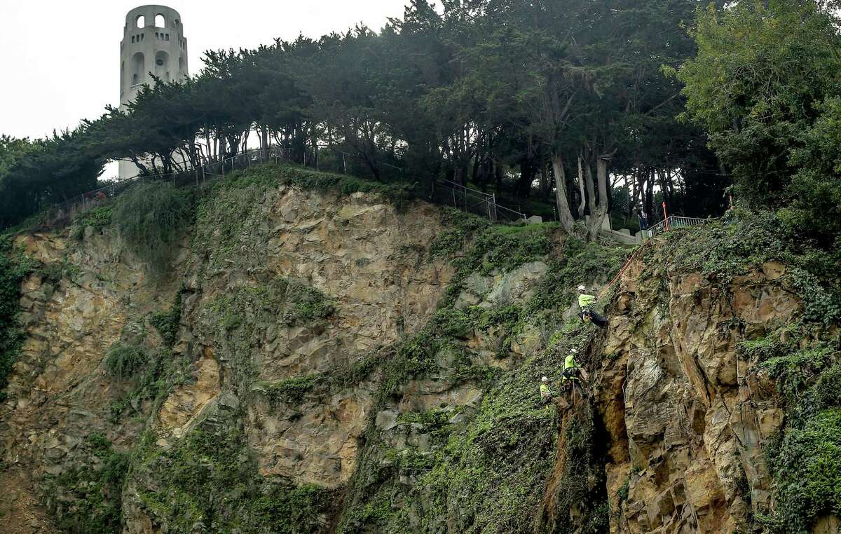 Workers hang from ropes to remove crumbling rock and plants as they start stabilizing the hill’s north slope.