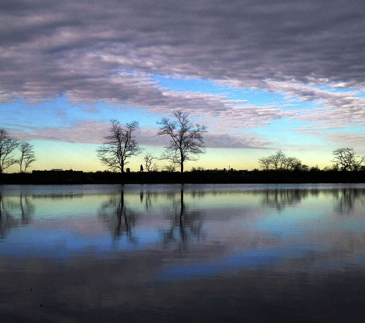 The silhouette of a walker is visible between a pair of trees on the horizon and is reflected, along with the surroundings, in Eagle Pond at Greenwich Point during sunset, Monday, March 17, 2014.