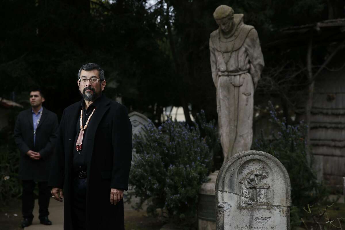 After news that Pope Francis will canonize Father Junipero Serra, Asst. curator of the Mission Dolores museum Vincent Medina with museum curator Andrew Galvan, both Ohlone Indians, stand in the Mission Dolores cemetery next to a statue of Junipero Serra on Thursday Jan. 15, 2015 in San Francisco, Calif. Galvan said their are 5,700 Indian buried in unmarked graves there. He hopes soon more will be memorialized.