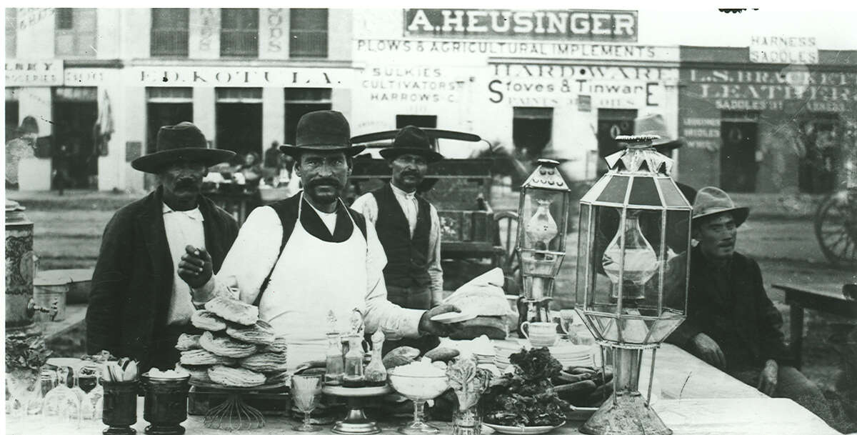 Men work in a chili stand on Military Plaza. Stores owned by Ed Kotula, A. Heusinger and L.S. Brackett, located on south side of plaza, are visible in background in this photo circa 1887-1890.