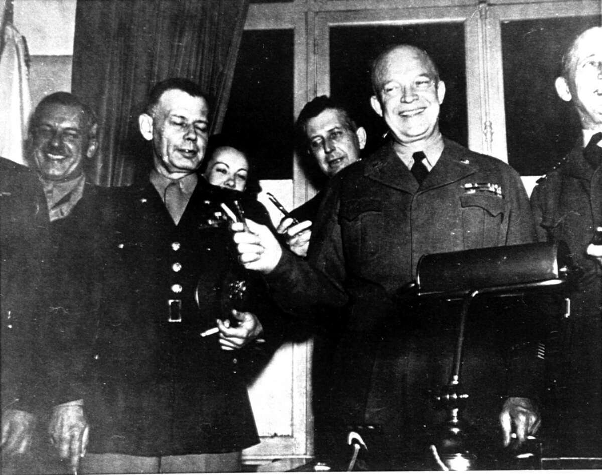 General Dwight D. Eisenhower holds up a V-for-Victory gesture with the two pens used by high ranking German officers in signing the surrender document at the school house in Reims, France on May 7, 1945. Kay Summersby, background left, Eisenhower's chauffeur-secretary, is seen peeking over General Walter Bedell Smith's shoulder. Her face was censored from the official photo taken by T/Sgt. Al Meserlin, Ike's personal photographer. Other officers are not identified. (AP Photo/Al Meserlin)