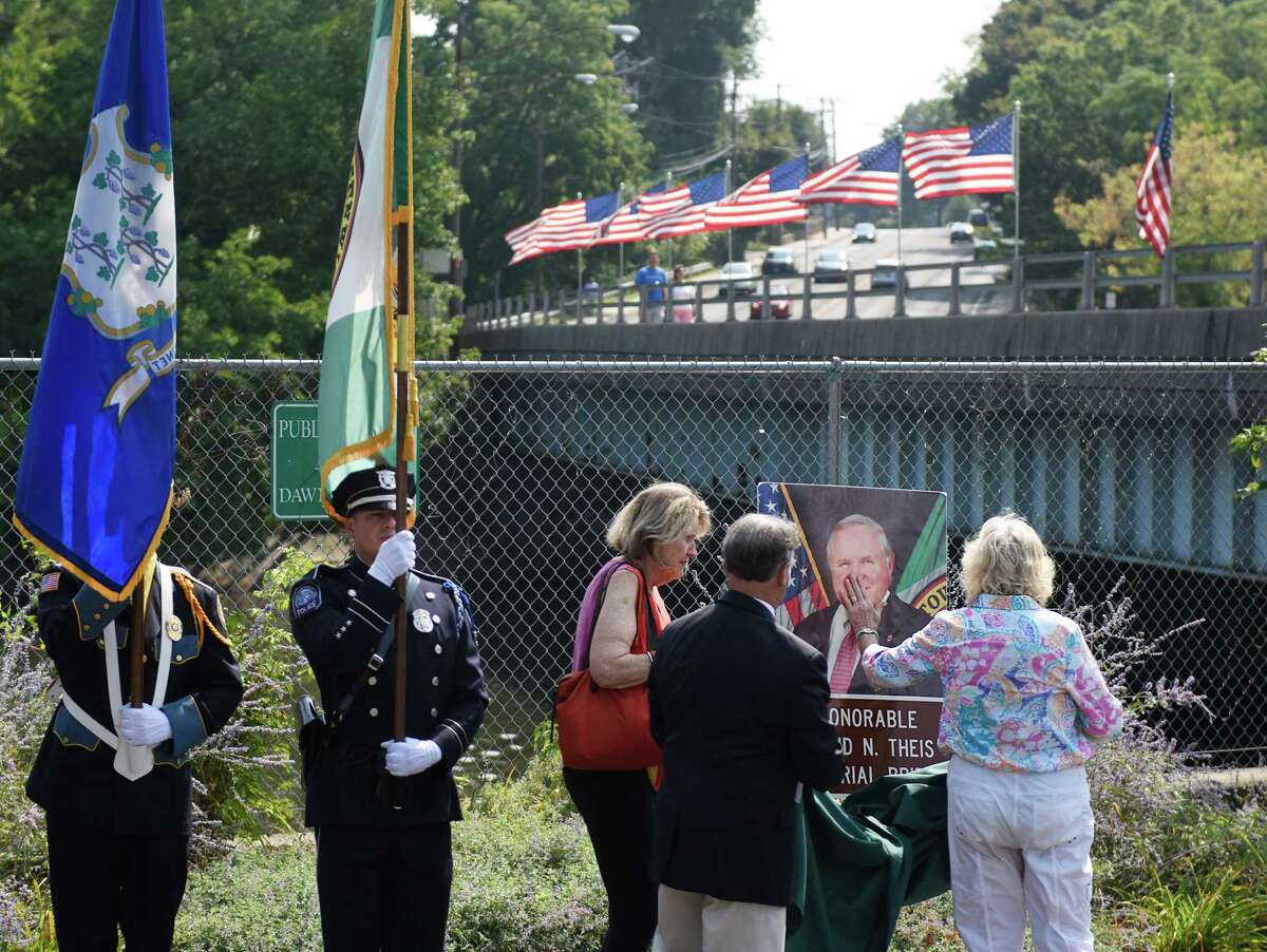 Dave Theis' longtime partner Kerrin Coyle kisses her hand and touches it to a photo of Theis, standing beside Theis' sister Ginny Theis, State Rep. Fred Camillo, center, and Cos Cob Volunteer Firefighter Fred Pugni after a photograph of Theis and bridge signage were revealed in front of the bridge during the bridge renaming ceremony for the late town Selectman David Theis at the Route 1 bridge spanning the Mianus River between the Cos Cob and Riverside sections of Greenwich, Conn. Sunday, Aug. 30, 2015. The bridge was officially renamed The Honorable David N. Theis Memorial Bridge on what would have been Theis' 66th birthday. Theis, who died suddenly last December, worked to put up American flags on the bridge every summer to pay tribute to America and its armed forces.