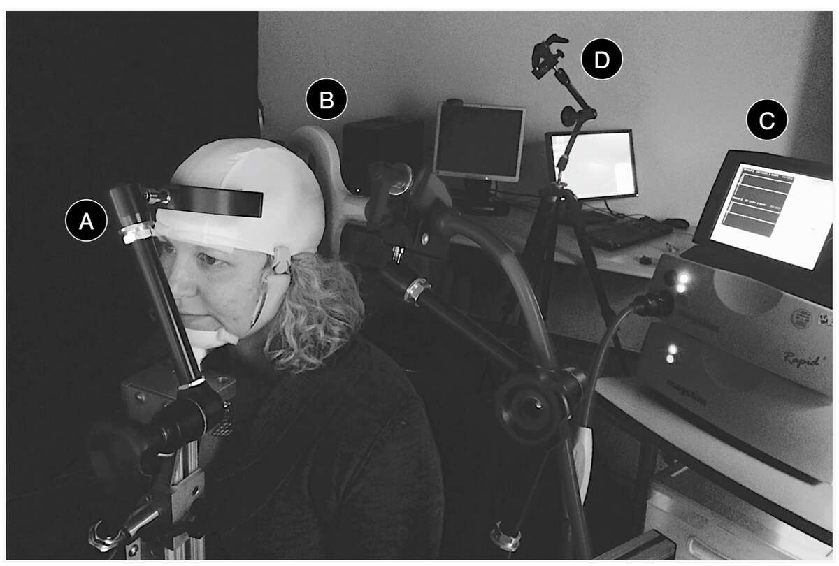 PLOS ONE caption: TMS Components of the BBI for the Inquirer. During the experiment, the inquirer sat in front of a computer screen on a BrainSight chair, with his or her head kept in place by a two-pronged head rest (A). A figure-of-8 TMS coil (B), connected to a MagStim Super Rapid2 stimulator (C) was positioned over the inquirer’s occipital lobe to deliver visual stimulation in accordance with the respondent’s answer. The head position and the coil position were carefully checked with the aid of a laser pointer (D). (Image from BBI research report in PLOS ONE)