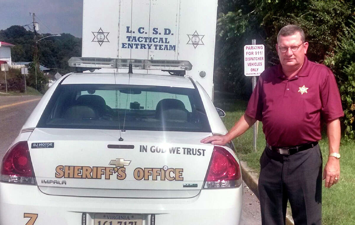 Lee County, Va., Sheriff Gary Parsons stands next to a patrol car that displays an "In God We Trust" decal on Friday, Sept. 4, 2015. Parsons said his office spent a total of $50 to have the decals added to about 25 vehicles. He said many people feel their belief system is being trampled and that adding the phrase is a way of pushing back. But a watchdog group says the decals amount to an illegal government endorsement of religion. (Rex Bailey/Lee County Sheriff's Office via AP)
