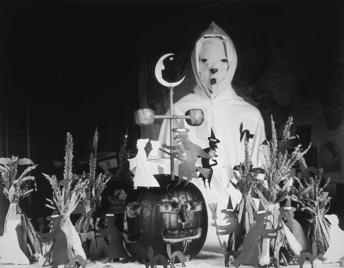 circa 1905: View of a person, possibly a teacher, wearing a ghost costume behind a lunch table with Halloween decorations in a rural schoolhouse. The display consists of a carved pumpkin, cutouts of witches and black cats, and haystacks. 