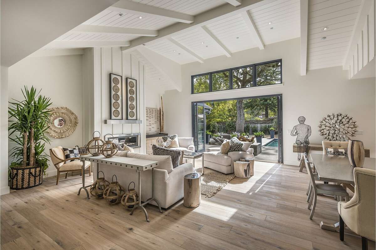 A vaulted, beamed ceiling towers above a great room with white oak flooring, a gas fireplace and accordion-style doors opening to the backyard.