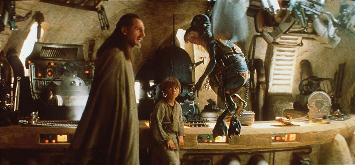 Jedi Master Qui-Gon Jinn (Liam Neeson) talks to junk dealer Watto about buying an uncommon spacecraft part as young Anakin Skywalker (Jake Lloyd) looks on in "Star Wars Episode I: The Phantom Menace." 