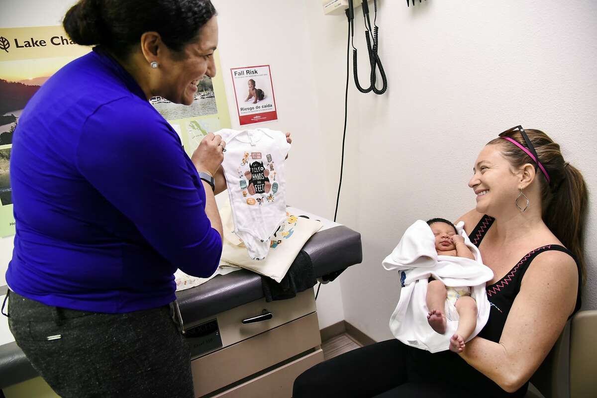 Dr. Dayna Long, left, holds up a onesie for 7 day old Amia Kelleher Taylor as she speaks with her mother Kimberly Taylor about the Opportunity Institute's "Talk, Read, Sing" program, during Kimberly's checkup visit to UCSF Benioff Children's Hospital Oakland Primary Care Clinic in Oakland, CA Tuesday, February 16, 2016.