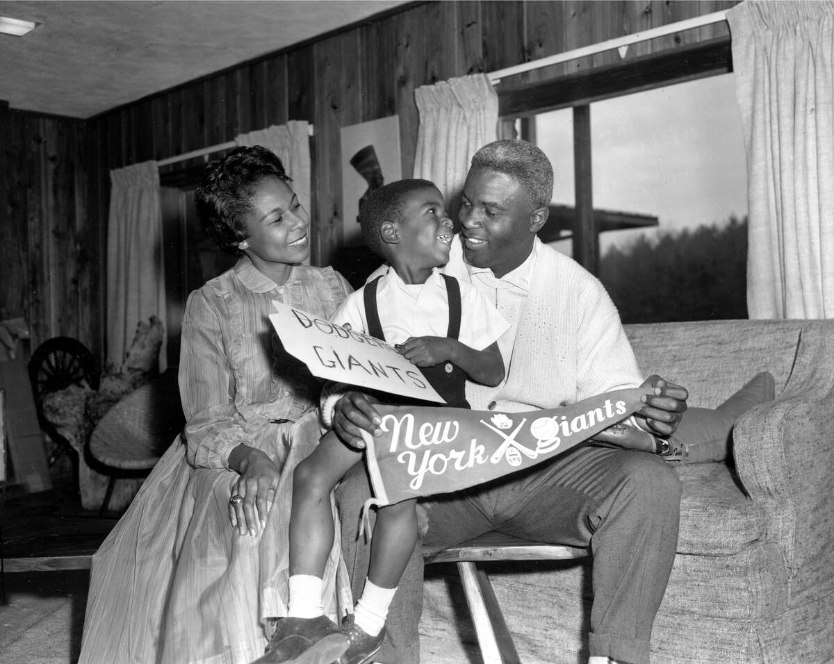Jackie Robinson, Brooklyn Dodgers star since breaking into Major League baseball in 1947, is shown with his wife, Rachel, and their four-year-old son, Jackie Jr., in their home in Stamford, Conn., Dec. 13, 1956. Robinson, 37, holds a banner for the rival New York Giants baseball club to which he was traded for relief pitcher Dick Littlefield and $35,000 in cash. Robinson retired in January, voiding the trade. (AP Photo/John Lindsay)