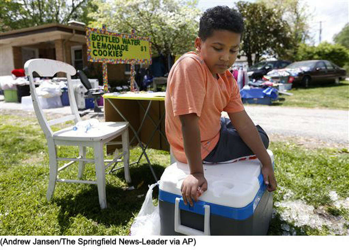 In this April 22, 2016 photo, Tristan Jacobson sits on a water cooler in front of his lemonade stand outside his home in Springfield, Mo. Tristan has been living with with Donnie and Jimmy Davis, who have been Tristan's kinship guardians. They have been holding a yard sale and set up a lemonade stand to raise money for his adoption. (Andrew Jansen/The Springfield News-Leader via AP) NO SALES; MANDATORY CREDIT