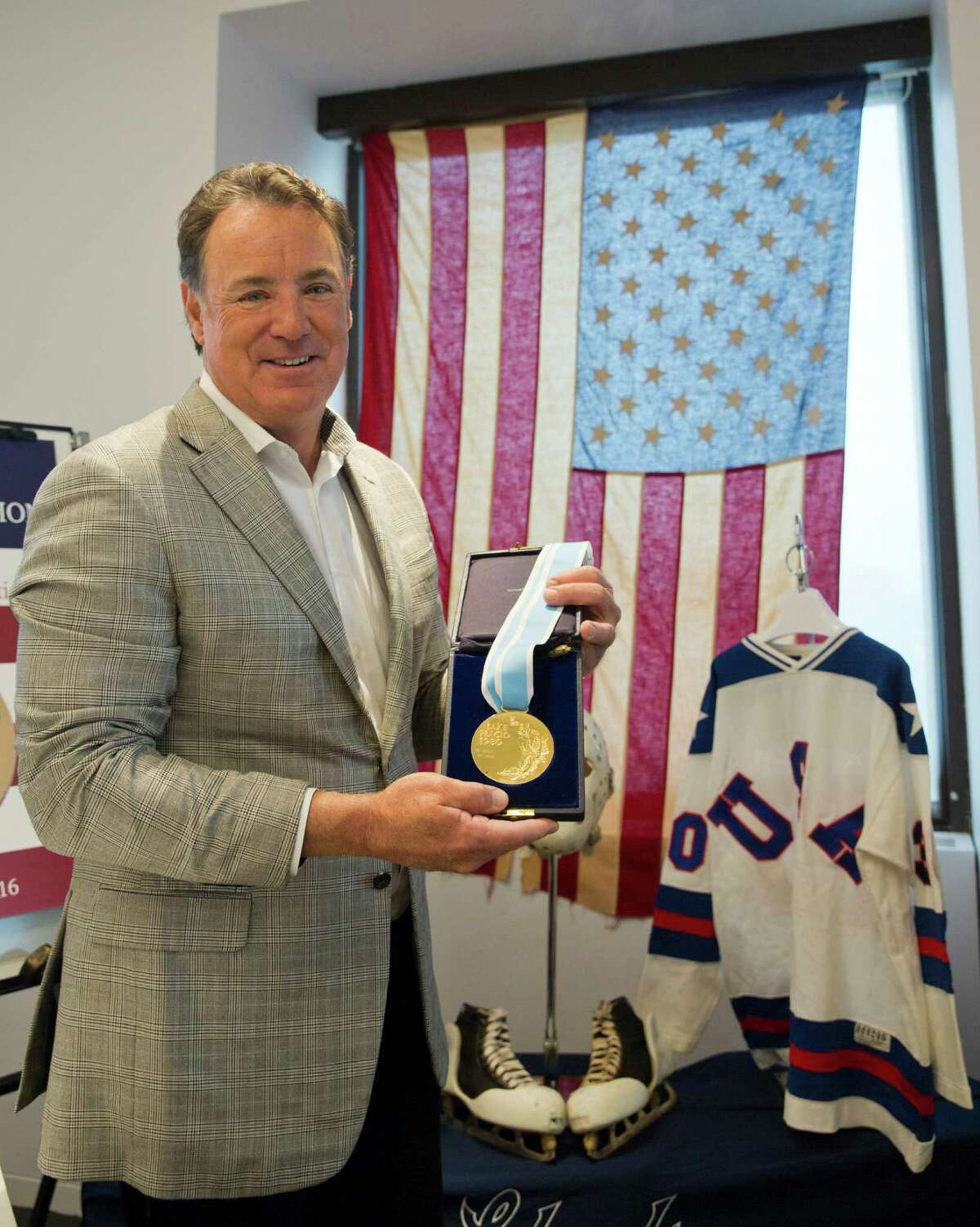 Jim Craig, the hockey goaltender who helped the U.S. win a miraculous gold medal at the 1980 winter Olympics, is framed by a display featuring the jerseys, skates and goalie equipment he wore in the Soviet and Finland games as well as the iconic American flag that was draped over his shoulders after the gold medal win, as he poses for a photo with the his Olympic Gold medal, Tuesday, May 24, 2016, in New York. Craig is now auctioning off the items. (AP Photo/Mary Altaffer) ORG XMIT: NYMA102