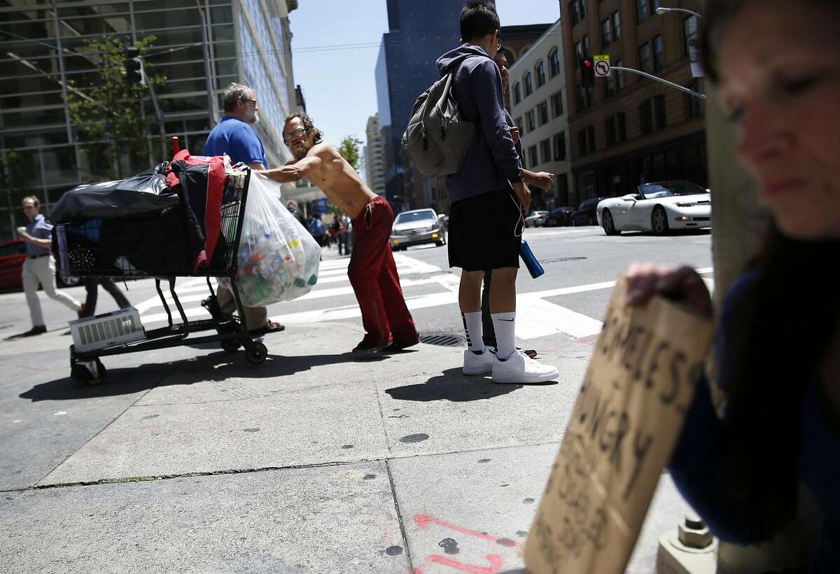 A man pushes a grocery cart across Second Street while Christine Boyer (right) panhandles on the corner of Mission and Second Street on Friday, May, 27, 2016 in San Francisco, California.