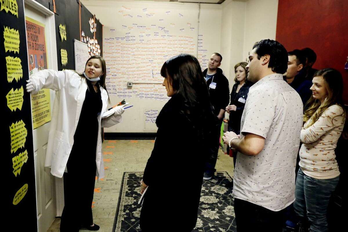 In a photo from Friday, Jan. 23, 2015 in Detroit, hostess Cassandra Maniak, left, explains the rules of the interactive theater to a group. Called “Trapped in a Room With a Zombie,” up to a dozen people are locked in a room for an hour alongside a zombie that’s chained to a wall. Every five minutes, a buzzer sounds, and the zombie is given another foot of chain, allowing the undead creature to scamper even closer to its next meal. If participants don’t solve a series of puzzles within the 60 minutes, the zombie “eats” everyone in the group. (AP Photo/Carlos Osorio)