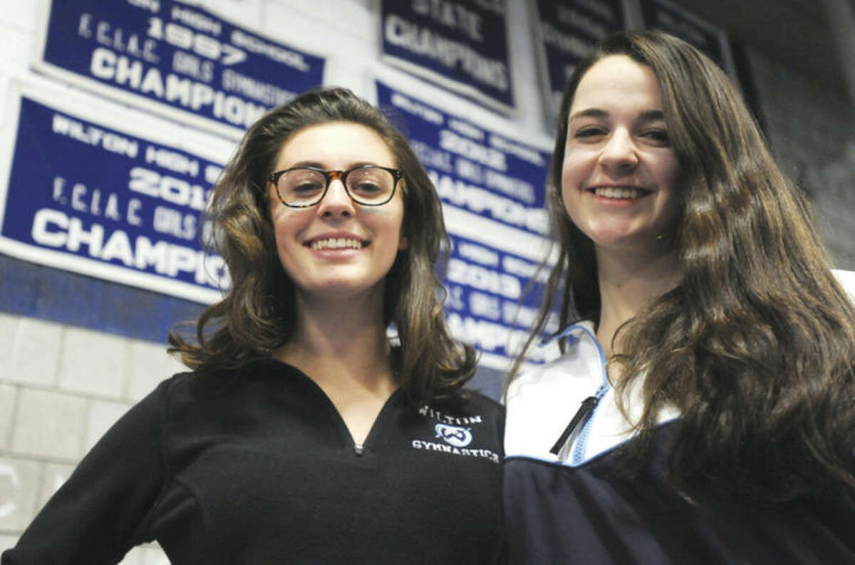 Hour photo/John Nash Wilton's Annie Saltarelli, left, and Meghan Graham, the co-captains of the Warriors gymnastics team, stand under the three previous banners the program has raised as three-time defending FCIAC champions. The Warriors are seeking their fourth straight title on Saturday.