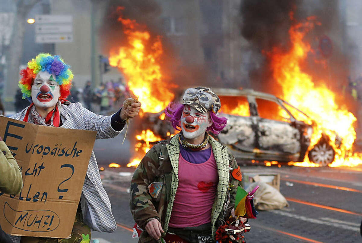 Demonstrators dressed as clowns pass by a burning police car Wednesday, March 18, 2015 in Frankfurt, Germany. Blockupy activists try to blockade the new headquarters of the ECB to protest against government austerity and capitalism. (AP Photo/Michael Probst)