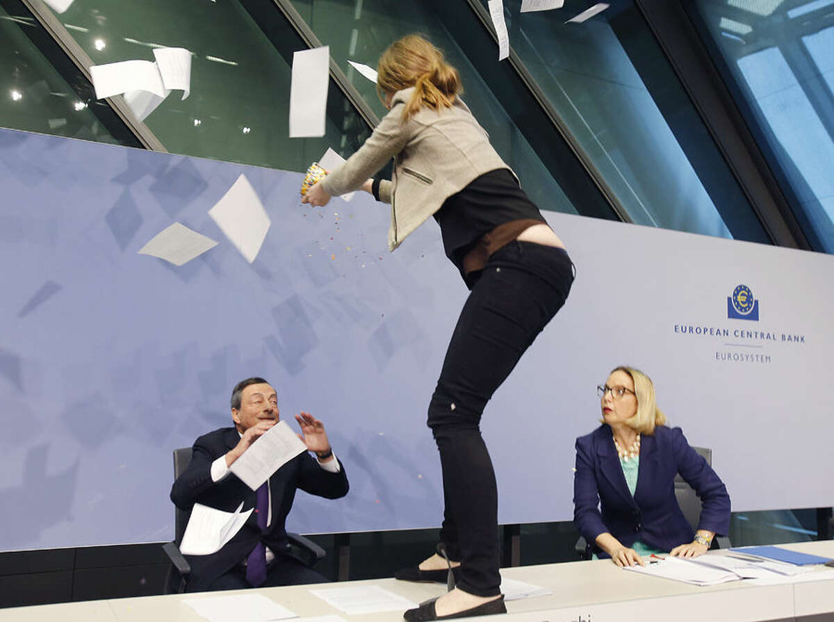 An activist stands on the table of the podium throwing paper at ECB President Mario Draghi, left, as Christine Graeff, Director General of Communications, looks on during a press conference of the European Central Bank, ECB, in Frankfurt, Germany, Wednesday, April 15, 2015. (AP Photo/Michael Probst)