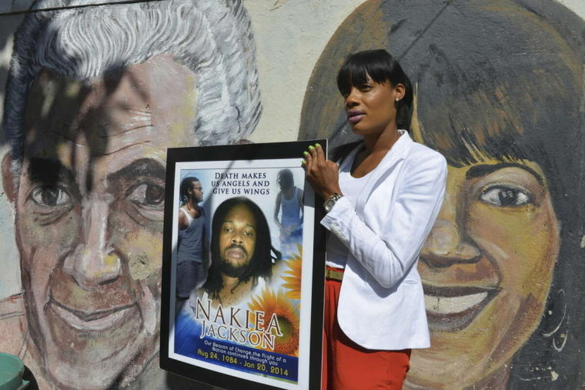 In this April 16, 2014, photo, Shackelia Jackson-Thomas holds a poster showing her brother, Nakiea Jackson, just outside his small ?“cookshop?” in the Orange Villa section of Jamaica?’s capital of Kingston. Earlier this year, the 27-year-old cook was shot dead by a policeman as pots of curried goat simmered on the stove of his kitchen, the exterior of which is decorated with murals of past and present political leaders. The patrolman who shot him is one of 27 police officers now facing charges of murder brought by an investigative commission probing allegations against police and soldiers. Jamaica?’s security forces have long been accused of indiscriminate shootings and unlawful killings. (AP Photo/David McFadden)