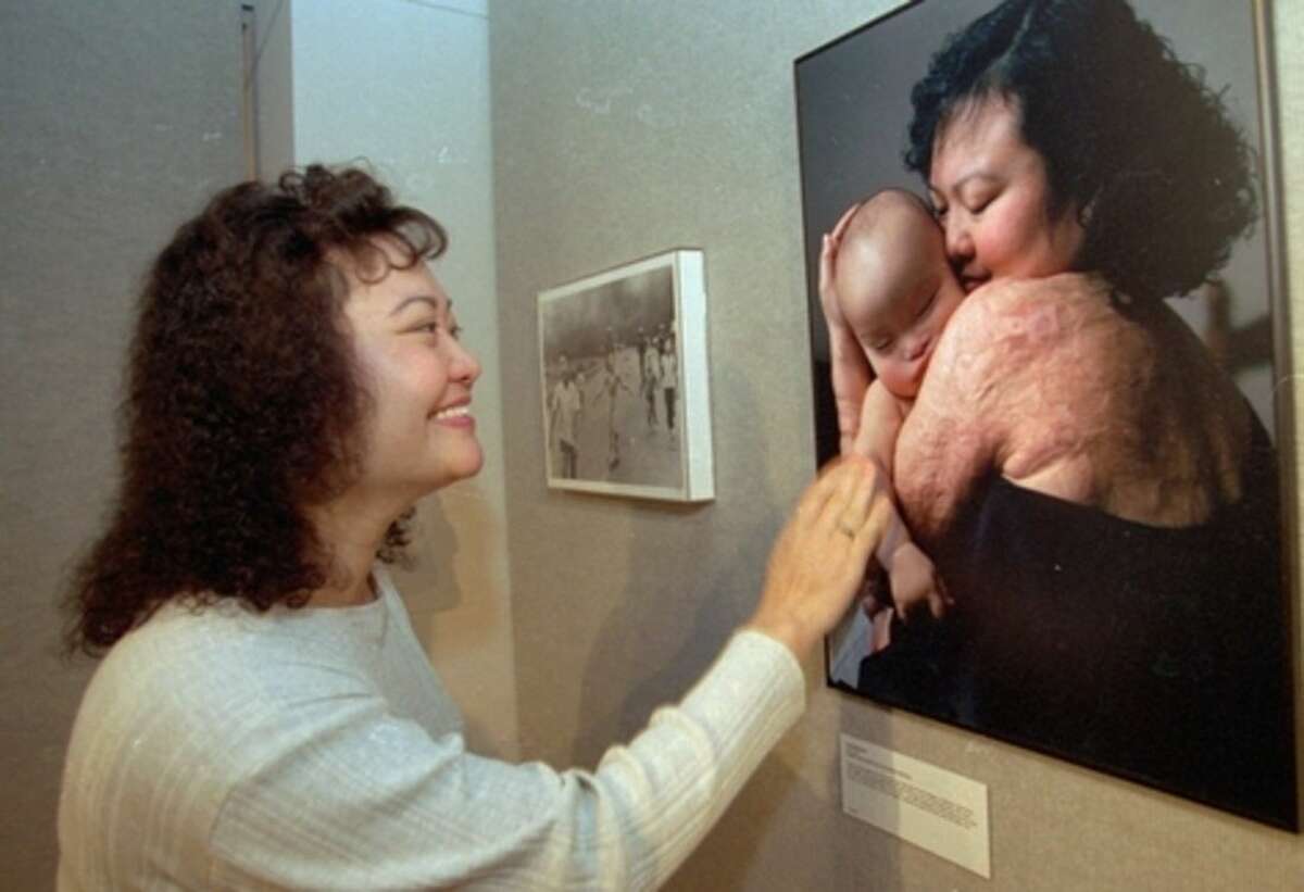FILE - In this Oct. 26, 1996, file photo, Kim Phuc, the victim of a napalm air strike in Vietnam in 1972, looks at a photo of herself holding her sleeping son, her back still bearing scars from the napalm burns, during a visit to the photography exhibit, "Eyewitness 1996," at the Museum of Tolerance in Los Angeles. The Pulitzer Prize-winning photo of Phan fleeing after the attack, by Nick Ut, is at left. (AP Photo/Nick Ut, File)