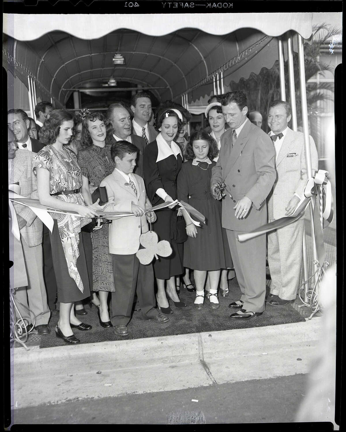 03/17/1949 - Ribbon cutting ceremonies at the grand opening of the Shamrock Hotel. two of Glenn McCarthy's daughters (not sure of order), Mary Margaret McCarthy and Glenna Lee McCarthy are on the left; young Glenn McCarthy Jr.; behind him are entertainer Pat O'Brien and actor Robert Paige; actress Dorothy Lamour; Glenn McCarthy's youngest daughter, Faustine McCarthy; Mrs. Glenn (Faustine) McCarthy; Glenn H. McCarthy holding the scissors.
