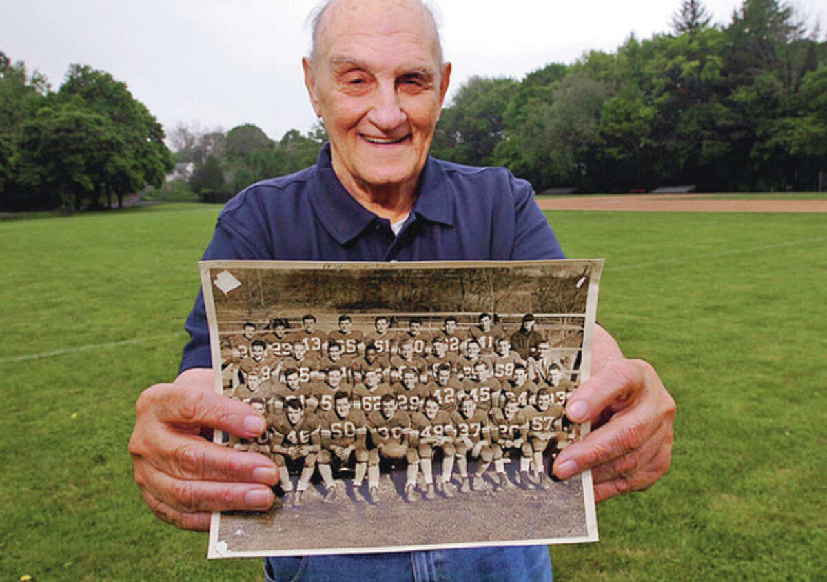Hour photo / Erik Trautmann Pat Nardi stands on the field behind City Hall where he once played as he proudly displays a photo of the 1951 Norwalk High School football team that upset unbeaten and top-ranked Danbury 19-14 on Thanksgiving morning. Nardi, the starting right guard and a two-way player on that team, is pictured in the front row, fourth from the right, wearing No. 49.