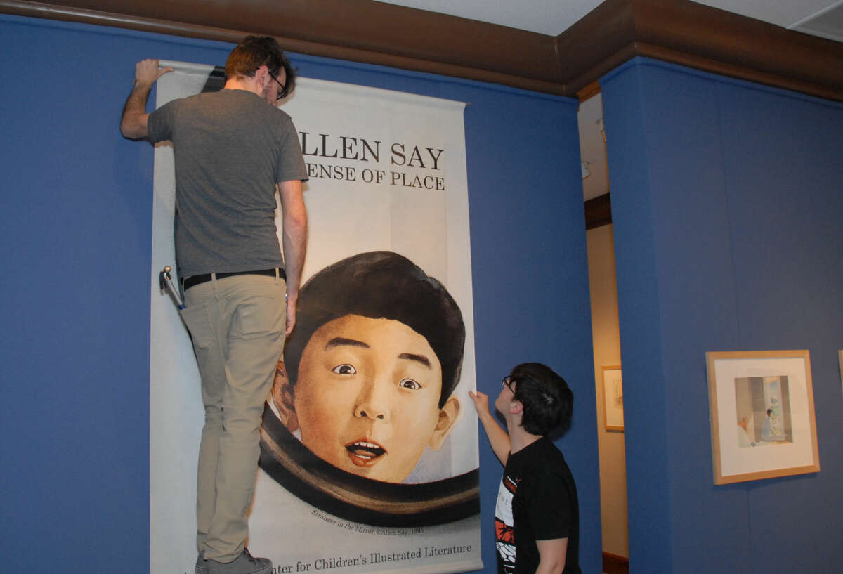 Wayland art students Jon Riddle, left, and Justus Brozek hang a banner welcoming visitors to the “Journey of Memory: Works of Allen Say” exhibit on display in the Abraham Art Gallery.