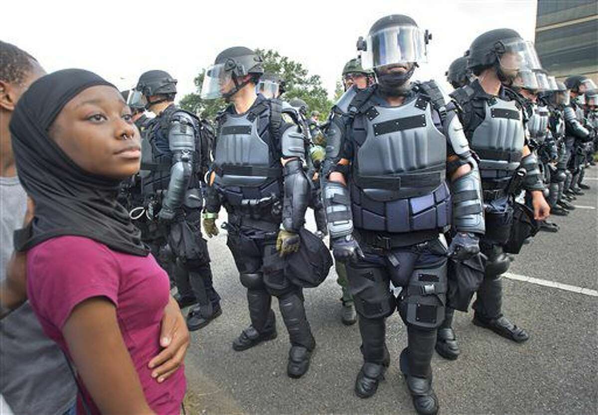 In this Saturday, July 9. 2016, photo, a protester awaits allowed safe passage across a highway previously blocked by protesters, as law enforcement officers wearing riot gear stand in formation to control the road in Baton Rouge, La., during demonstrations about the fatal shooting by police of Alton Sterling in Baton Rouge. The police department has come under criticism for the tactics it's employed to deal with protesters, using riot police and military-style vehicles on the streets of the capital city. Over a three-day period, police arrested about 200 protesters. Police arrested three suspects and were seeking a possible fourth suspect accused of stealing several handguns as part of what authorities Tuesday described as "substantial, credible threat" to harm police officers in the Baton Rouge area. "We have been questioned repeatedly over the last several days about our show of force and why we have the tactics that we have," Baton Rouge Police Chief Carl Dabadie said, "Well, this is the reason, because we had credible threats against the lives of law enforcement in this city." (Travis Sptradling/The Advocate via AP)