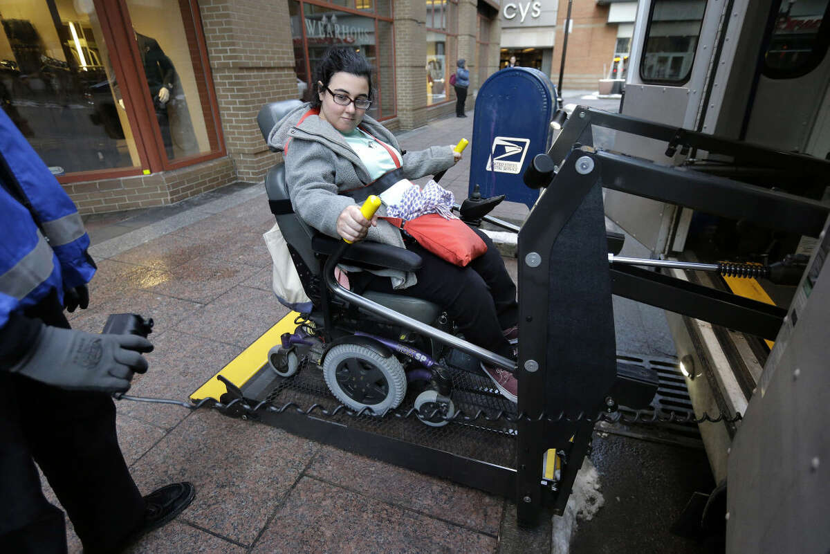 In this Tuesday, April 5, 2016 photo, Sarah Kaplan, of Lynn, Mass., rides an elevator to board a bus outside her place of work, in Boston. Transit systems in major U.S. cities are required by federal law to provide specialized services for disabled passengers. In Boston, the deficit-laden MBTA is considering ways of reducing service for the disabled and outsourcing more of it to taxis and ride-hailing services like Uber. (AP Photo/Steven Senne)