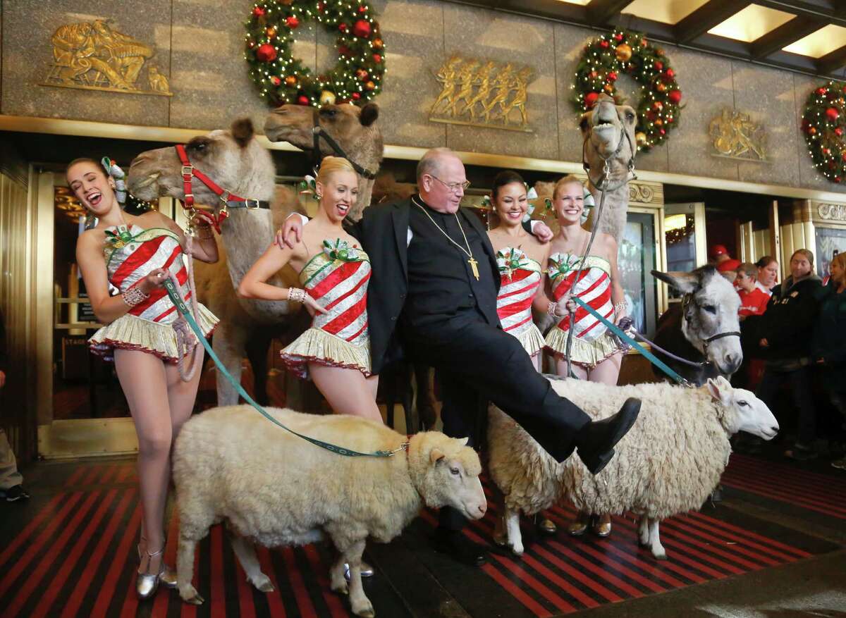 Cardinal Timothy Dolan, center, kicks his leg as he poses with members of the Radio City Rockettes, Jessica Davison, far left, Lauren Renck, second from left, Christine Sienicki, second from right, and Christine Walsh-Warren, far right, after he blessed camels, sheep and a donkey, Tuesday Nov. 1, 2016, in New York. The animals, arriving for their first day of rehearsal, will appear in the nativity scene of the "Christmas Spectacular Starring the Radio City Rockettes." (AP Photo/Bebeto Matthews) ORG XMIT: NYBM101