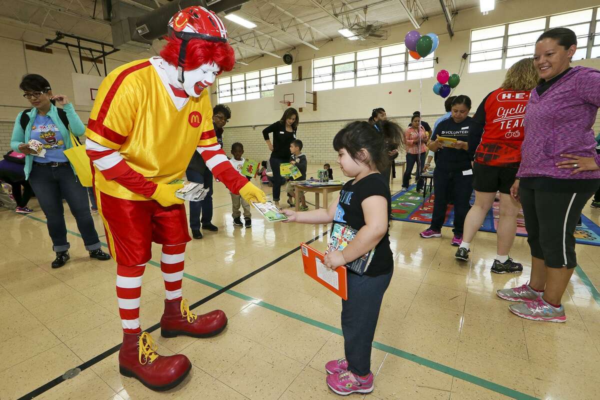 Ronald McDonald hands a Happy Meal Love Book to 4-year-old Faith Gonzales, as cyclists with Ride for Reading delivered books and provided tricycle safety tips to preschoolers at the Carroll Early Childhood Center. Approximately 40 volunteers from USAA and H.E.B participated in the organization's inaugural delivery in San Antonio, bringing 650 books to the children.