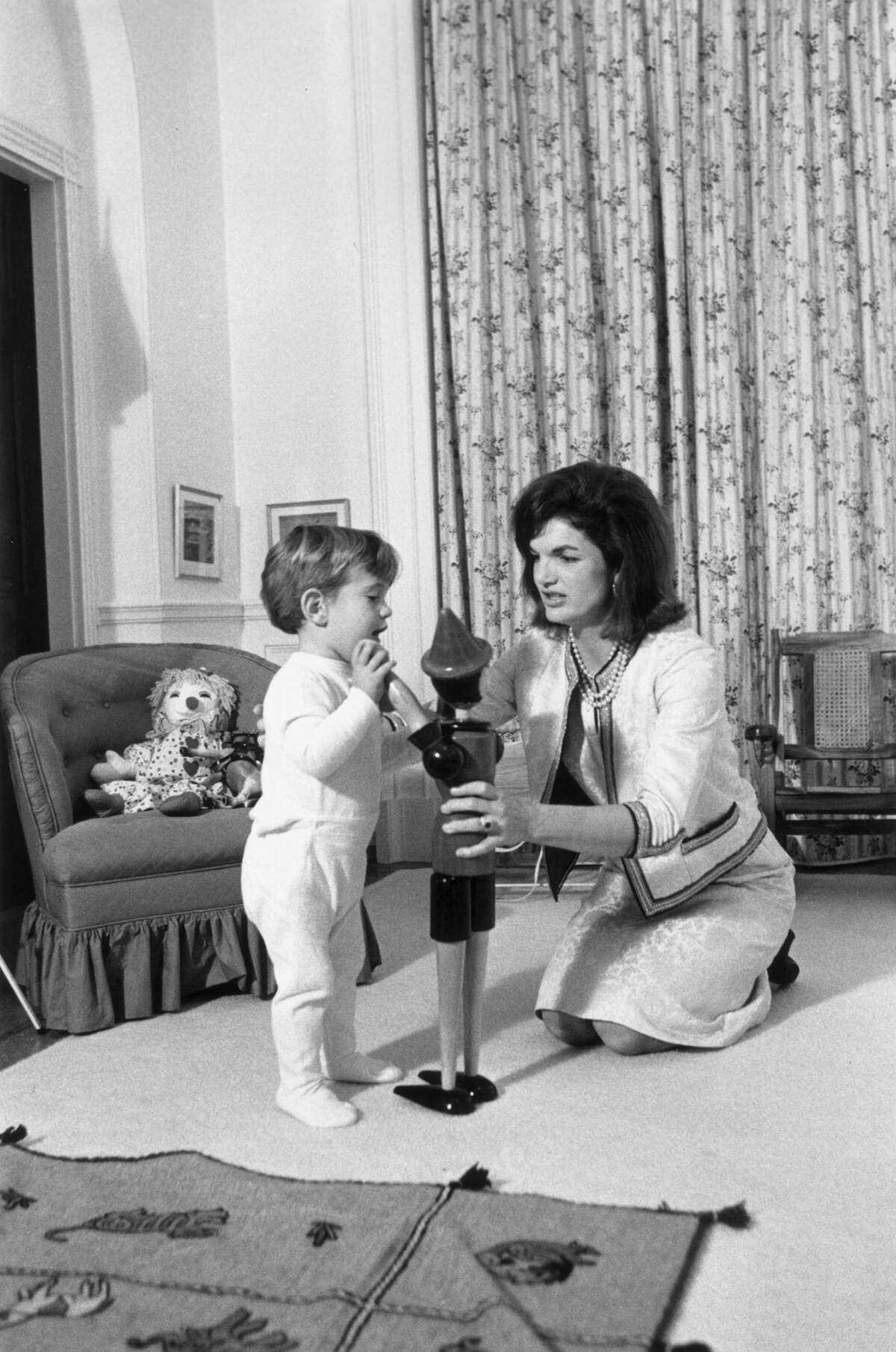 November 1962: American First Lady Jacqueline Bouvier Kennedy (1929 - 1994) kneels on the floor with her son, John F. Kennedy, Jr. (1960 - 1999), playing with a wooden toy figure, inside the White House, Washington, D.C. There is a rag doll on the chair behind them. (Photo by John F. Kennedy Library/John F. Kennedy Library/Getty Images)