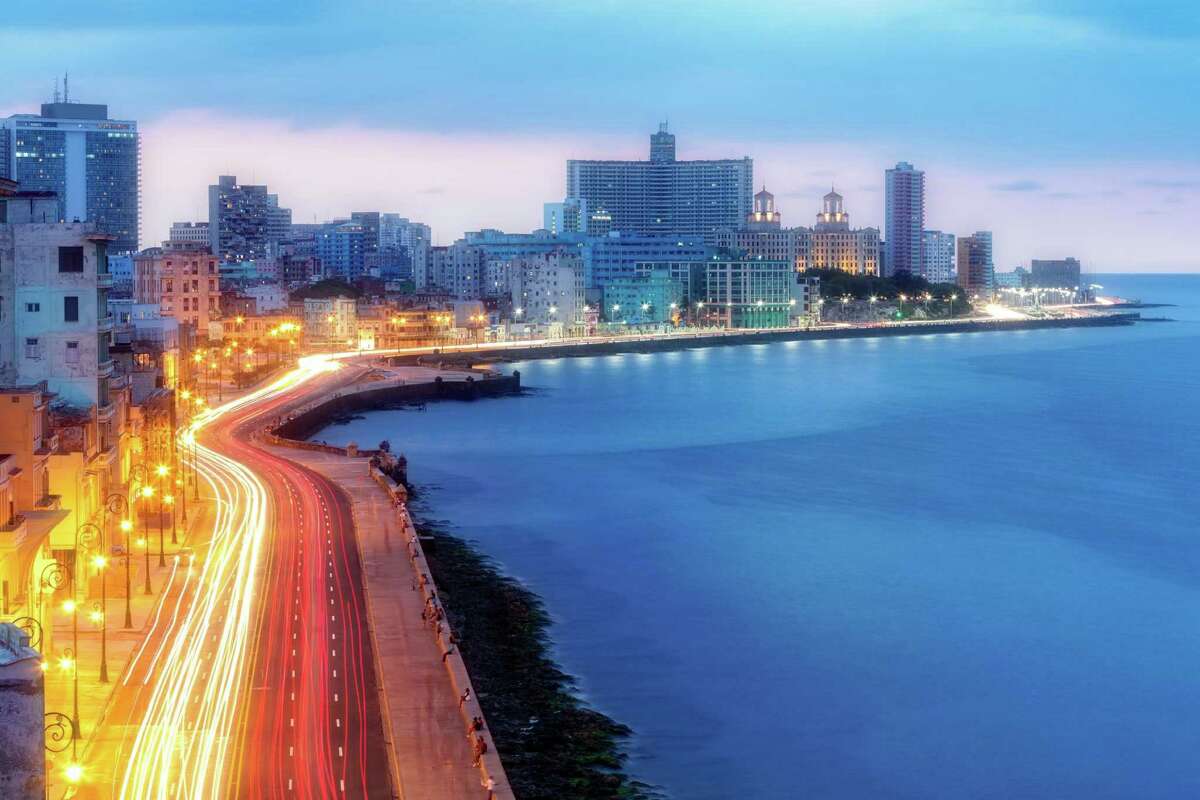 The Malecon seafront promenade at early morning with car light trails on the street, a very calm sea and the skyline of modern Havana with hotels is visible in the background.