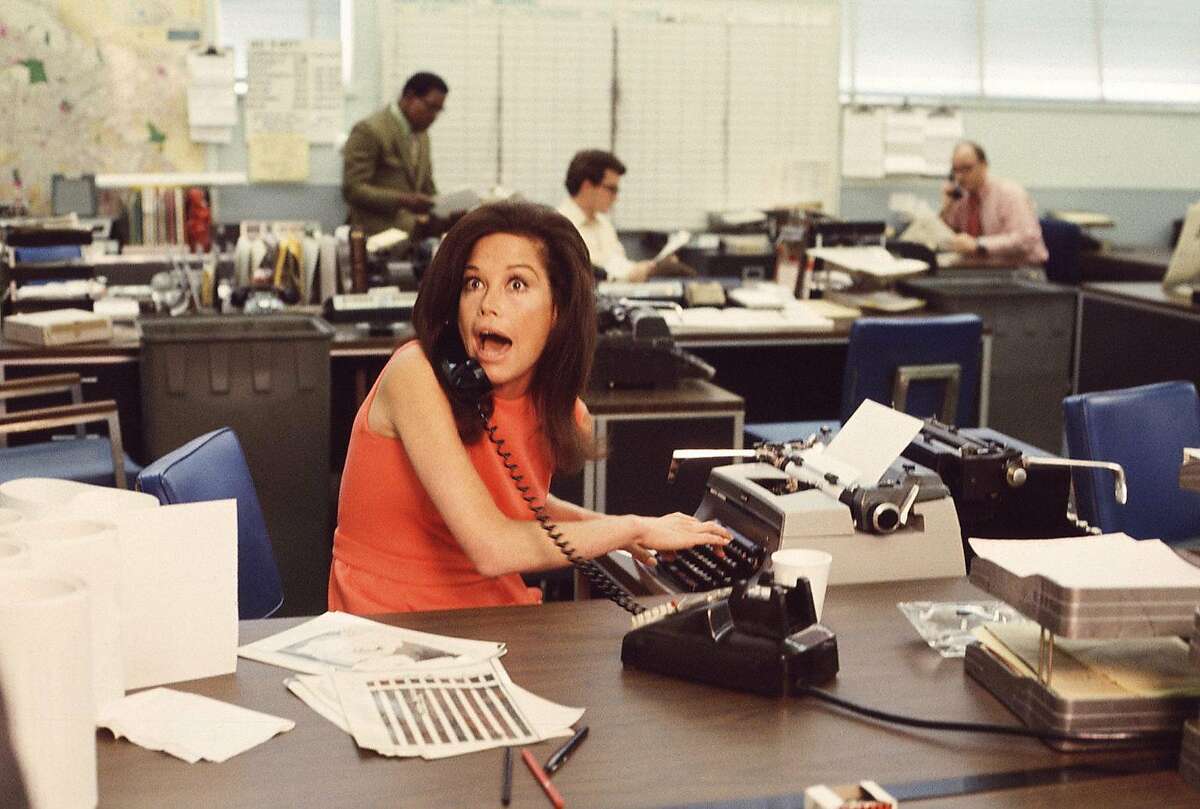 American actress Mary Tyler Moore mouths surprise on the telephone while simultaneously typing as others work in the background in a scene from 'The Mary Tyler Moore Show' (also known as 'Mary Tyler Moore'), Los Angeles, California, early 1970s. Moore wears a sleeveless orange dress as she sits behind her desk. (Photo by CBS Photo Archive/Getty Images)