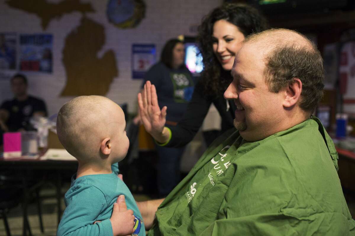 THEOPHIL SYSLO | For the Daily News Marc Thrush, paramedic with Midland EMS, rests his son Austin Thrush, 2, on his lap while Austin gives a high-five to Shannon Rose, employee at Lux The Salon, while participating in a head shaving fundraiser for cancer at The Boulevard Lounge on Sunday.