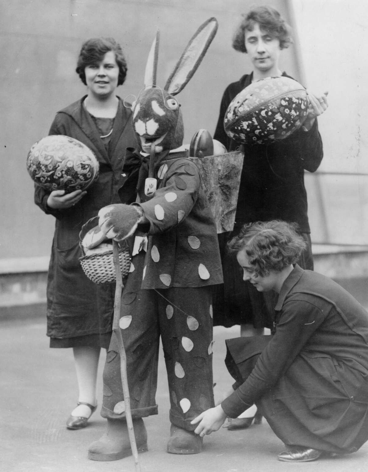 circa 1930: An Easter bunny carries easter eggs in a basket on his back while one woman adjusts his turn-ups and two others carry large easter eggs.