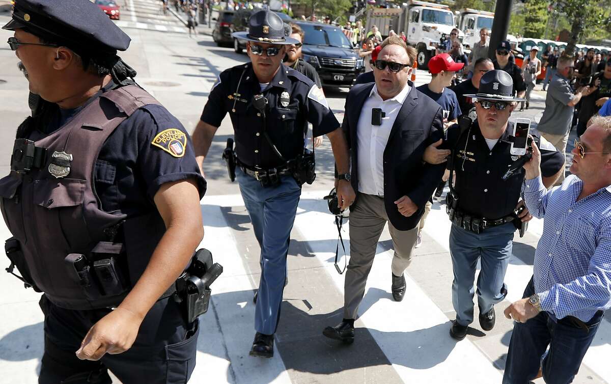 FILE - In this file photo made Tuesday, July 19, 2016, Alex Jones, center right, is escorted by police out of a crowd of protesters outside the Republican convention in Cleveland. A lawyer defending Jones, a the right-wing radio host and conspiracy theorist, in a child-custody dispute said Jones is a "performance artist" whose on-air persona differs from the private man. The Austin American-Statesman reported that Kelly Jones described her ex-husband at a recent pretrial hearing in Austin as "not a stable person." (AP Photo/John Minchillo, File)