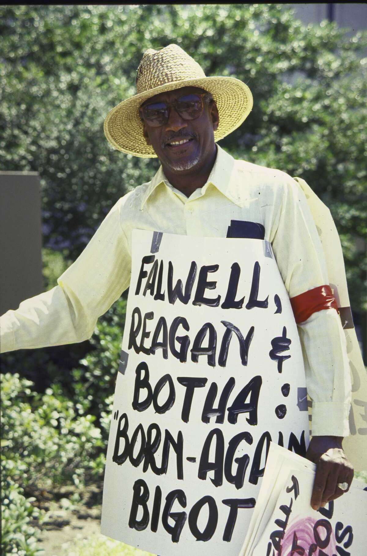 Counter-protestor wearing sign against apartheid, criticizing US Pres. Ronald W. Reagan regarding his South African policy of constructive engagement, along with the names of Moral Majority Leader Rev. Jerry Falwell and So. African Pres. Pieter Willem Botha, calling them Born Again Bigots during Falwell's anti-pornography demonstration.