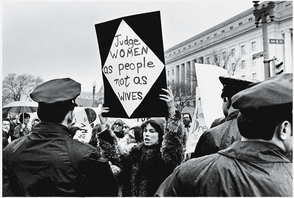 A young American woman holds up a sign as she protests for women's rights in front of the Federal Trade Commission headquarters while policemen look on during Richard Nixon's inauguration weekend, Washington, DC, January 18-21, 1969. Her sign reads 'Judge women as people not as wives.'