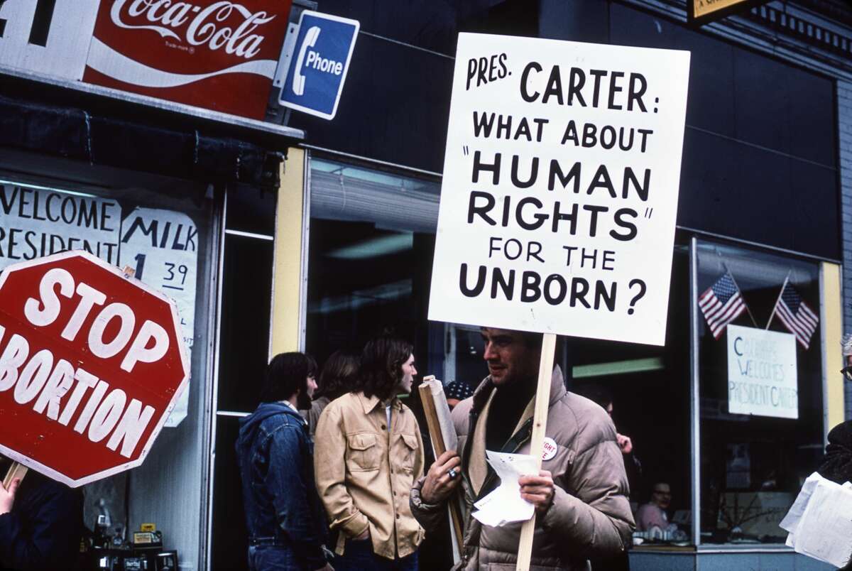 View of demonstrators on either side of the abortion debate stand on a sidewalk during a visit by US President Jimmy Carter, Clinton, Massachusetts, March 16, 1977. One man holds a sign that reads 'President Carter: What about human rights for the unborn?' while at left is a sign that reads 'Stop Abortion.'