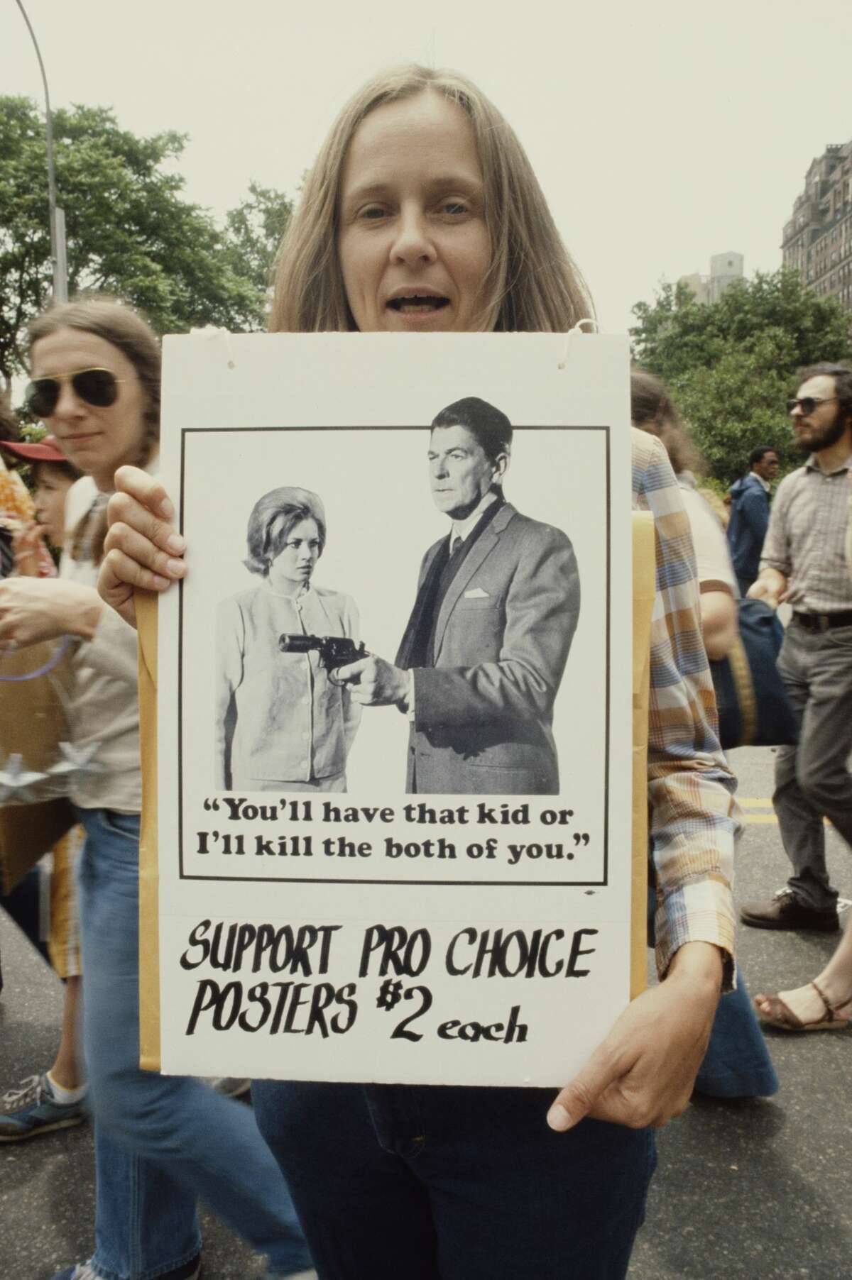 A woman selling pro-choice posters of US President Ronald Reagan threatening a woman with a handgun at a demonstration, circa 1987. The caption reads 'You'll have that kid or I'll kill the both of you'.
