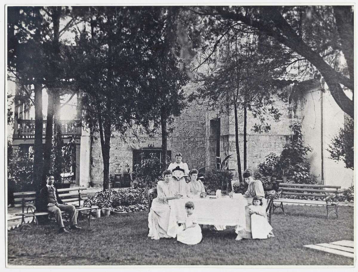 The facade and garden at the Anton Wulff House, looking southwest in 1892. Pictured are three of eleven children and two of the granddaughters of Anton Frederick and Paula Wulff. (Left to right) Maria Guadalupe Paulita Wulff, (maid not identified), Isabel (Lula) Wulff and Maria Wulff. Children in front of tea table are Anita and Linda Tyrrasch. The man on the bench at left is an unidentified friend. Black and white.