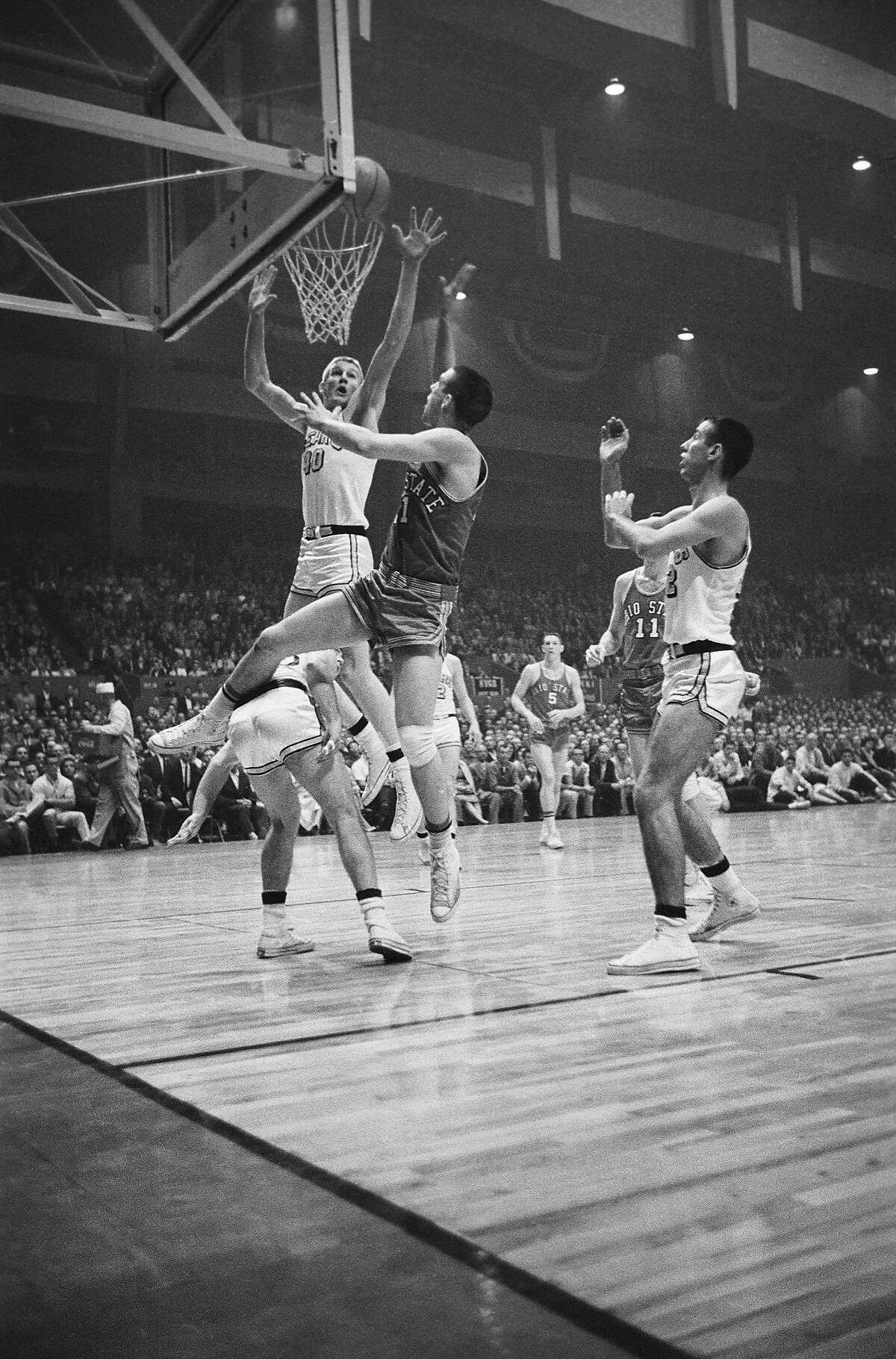 (Original Caption) With one leg draped over an unidentified player, Ohio State's Larry Sigfried (21) fires and scores from beneath the basket in the NCAA Championship game with California here on March 19th. Making a vain effort to block the shot is California's Darrall Imhoff, (40). At right is California's Iandy Gillis. Ohio State swamped California 75-55.