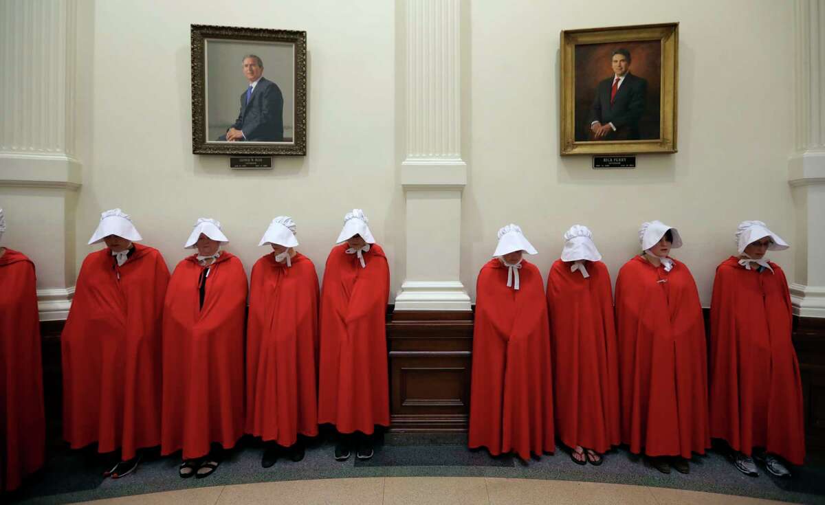 Women's rights activists dressed as characters from "The Handmaid's Tale" gather under portraits of former Texas governors George W. Bush and Rick Perry. The characters are based on a sci-fi novel and TV show about women forced to serve as reproductive vessels for infertile elites.