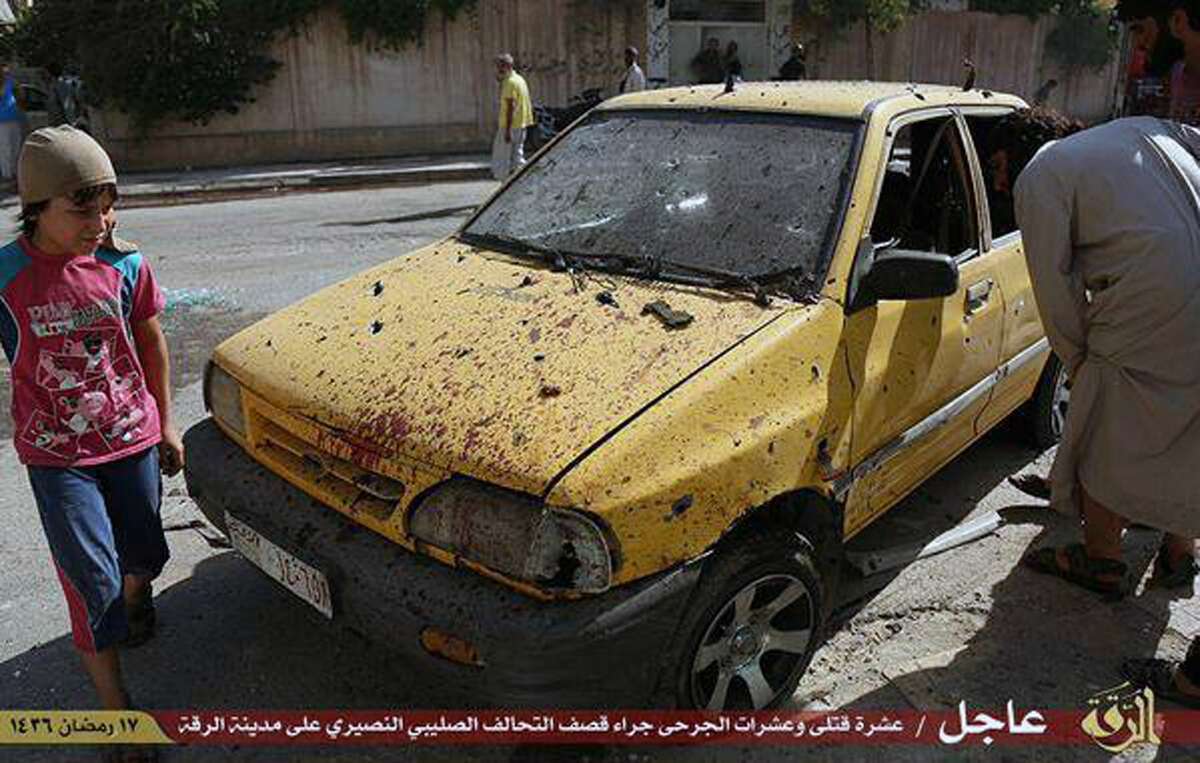 This July, 4, 2015 photo provided by a website of the Islamic State group, shows people inspecting a car purportedly damaged by U.S.-led airstrikes on Raqqa, Syria. A series of U.S.-led coalition airstrikes targeting the Islamic State group’s stronghold of Raqqa in eastern Syria has killed at least 10 militants and wounded many others. The airstrikes were confirmed by the coalition, Raqqa-base activists and the Islamic State group. Arabic on banner reads, “Raqqa Urgent / Ten killed and tens wounded by crusader (Christian)-Nusayri (Alawite) coalition on Raqqa city. 17 Ramadan 1436 Hijri.”