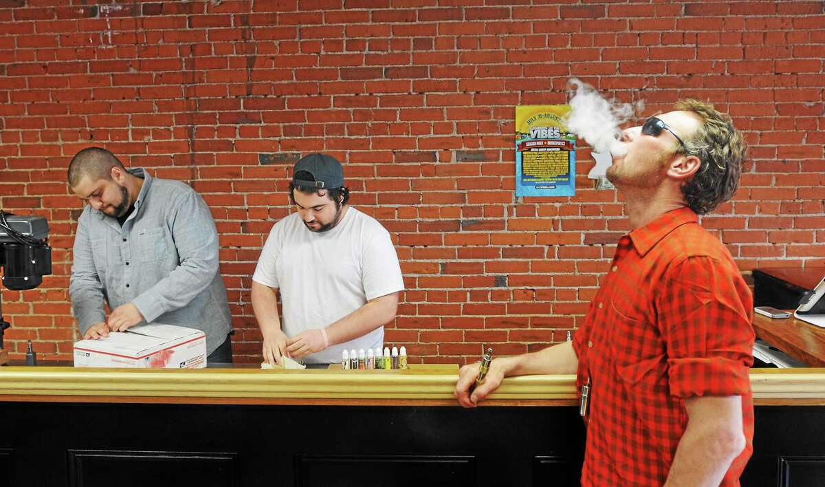 Customer Raymond Vickers of West Haven enjoys some “vape” as he hangs out in the lounge area of White Buffalo Vape shop. Employee Nick Anderson, left, and co-owner Noah Morgenstein organize a new shipment of products.