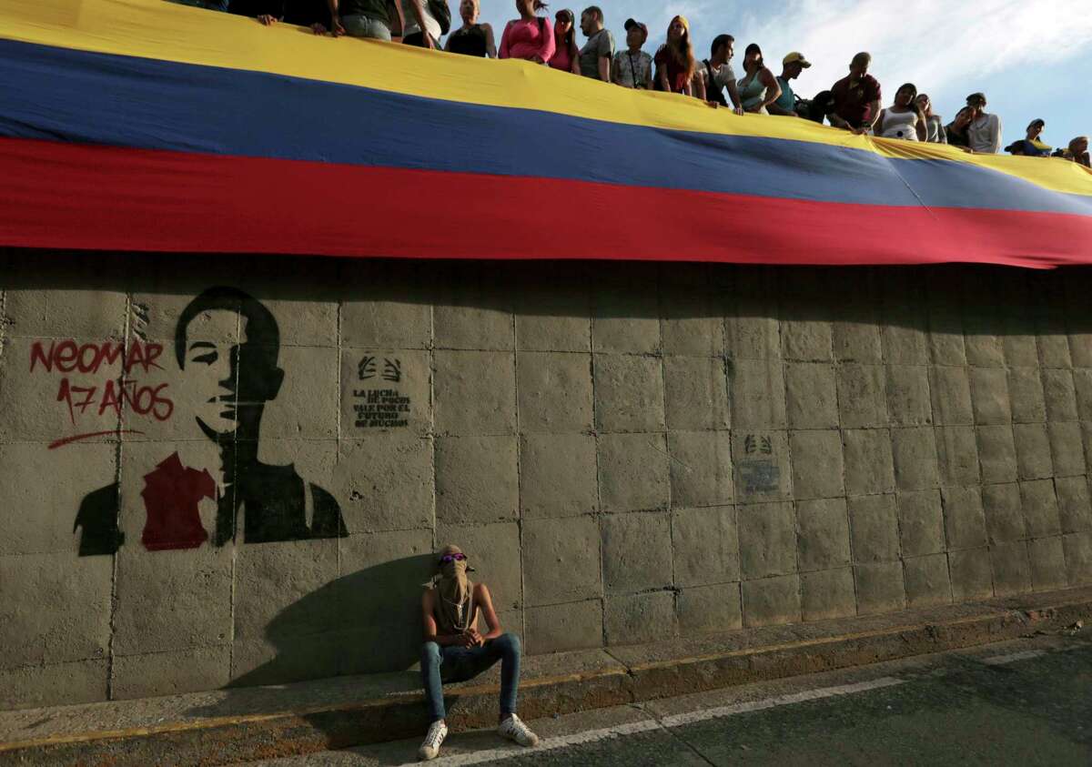 In this Monday, July 24, 2017 photo, a demonstrator sits near a spray-painted stencil of Neomar Lander during a tribute to those killed in the recent wave of protests against Venezuela's President Nicolas Maduro, in Caracas, Venezuela. An AP tally of official reports shows that the death toll in nearly 4 months of civil unrest in Venezuela reached 100 on Thursday, July 27, 2017. Most of the deaths reported by the countryÂ?’s chief prosecutor since anti-government protests began in April are of young men killed by gunfire. The count also includes looters, police allegedly attacked by protesters and civilians killed in accidents related to roadblocks set up during demonstrations. (AP Photo/Fernando Llano)
