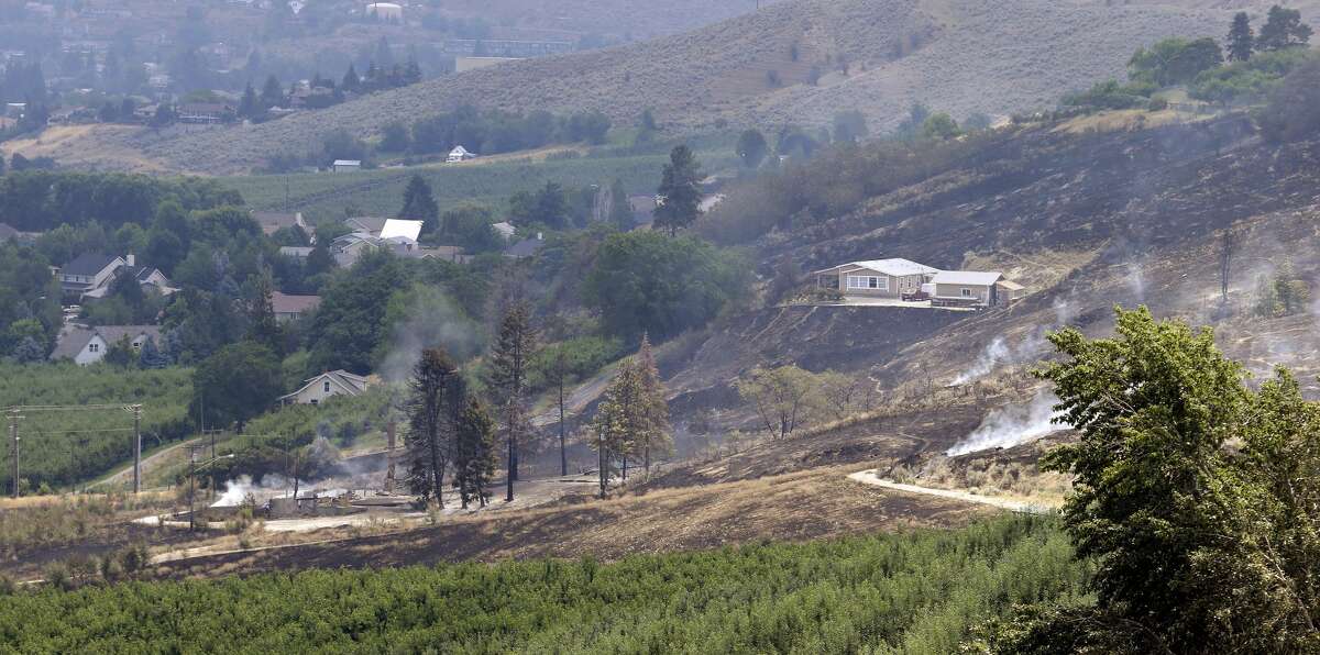 The foundation and chimneys from a destroyed home continue to smolder, lower left, in view of a home at right that survived despite the area around it being burned out from a wildfire that raced through the area the night before, Monday, June 29, 2015, in Wenatchee, Wash. The wildfires hit parts of central and eastern Washington over the weekend as the state is struggling with a severe drought, destroying dozens of structures and forcing hundreds to flee. (AP Photo/Elaine Thompson)
