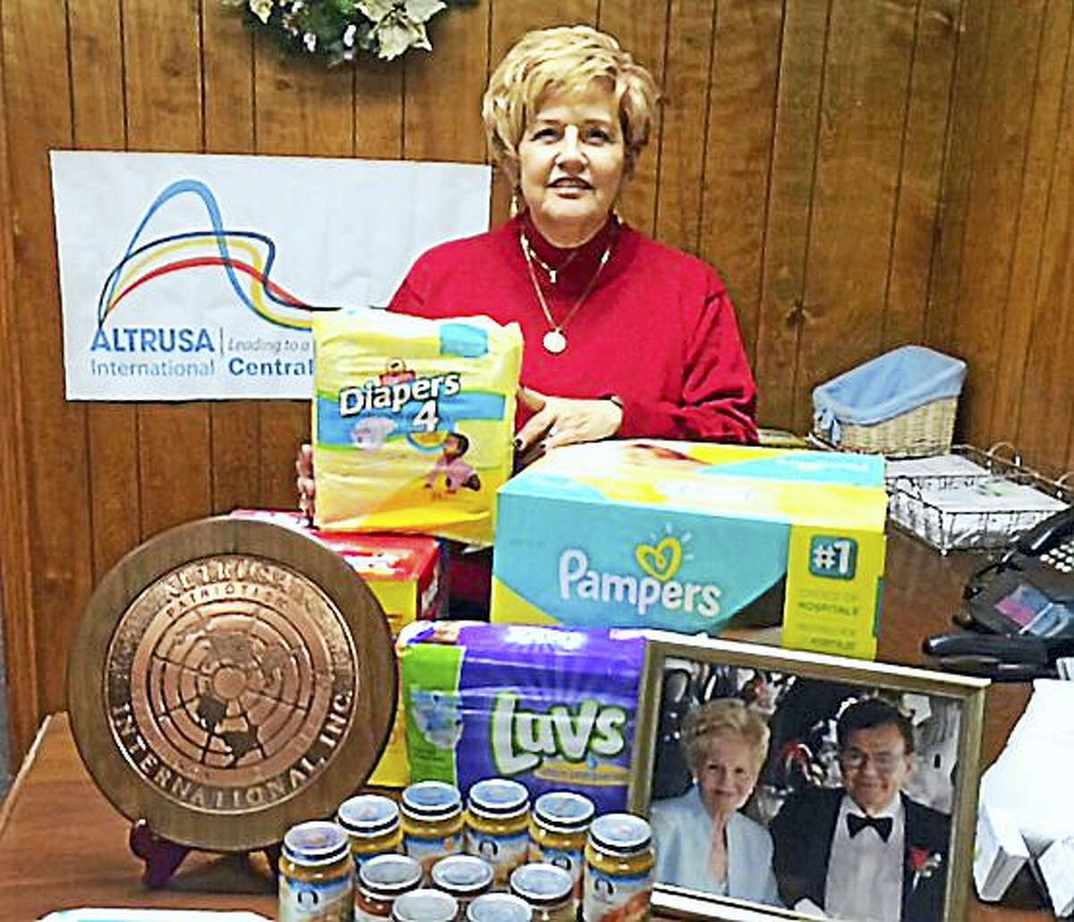 Kathleen Didato poses in front of an Altrusa banner, surrounded by donations for babies, and a photo of her parents, who inspire her work.